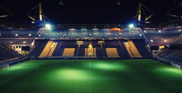 Ein Fußballfeld bei Nacht mit hellen Stadionlichtern, saftig grünem Rasen und einigen Zuschauern auf den Tribünen.
