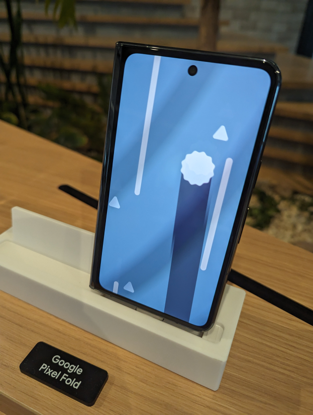 Ein Samsung Galaxy Fold 2 Smartphone mit gebogenem Display und Metallrahmen steht auf einem Tisch mit einer Namensschilder, umgeben von Pflanzen und einer Wand im Hintergrund.