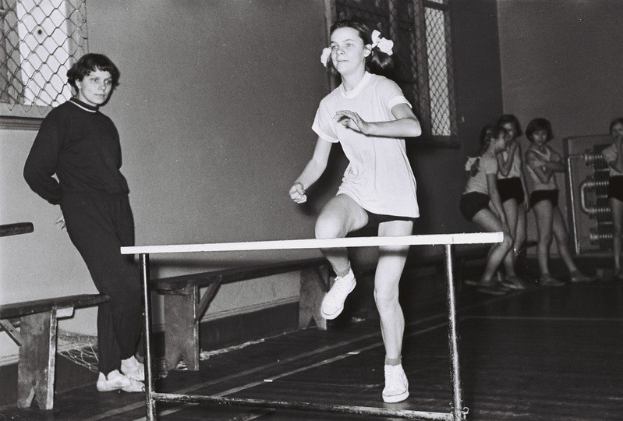 Schwarzes und weißes Foto einer Frau in der Luft, die über ein Hindernis in einer Turnhalle springt, mit Zuschauern und Fenstern im Hintergrund.