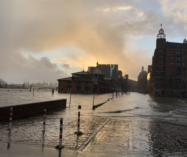 Überschwemmte Straße in Hamburg, Deutschland mit Wasser auf der Straße, Pfählen, Schildern, Gebäuden mit Fenstern, einer Brücke und einem bewölkten Himmel im Hintergrund.