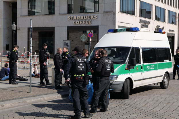 Eine Gruppe von Polizisten steht vor einem Starbucks-Café, rechts ist ein Van zu sehen und links ein paar Menschen; im Hintergrund ist ein Gebäude mit Fenstern, ein Schild, ein Laternenmast und ein Zaun zu erkennen.