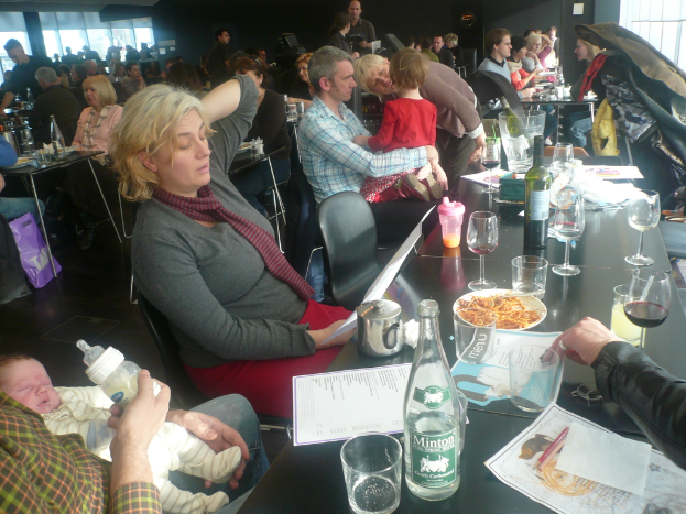Ein Innenblick in ein belebtes Restaurant mit Menschen an Tischen, die mit Gläsern, Tellern mit Essen, Getränkeflaschen, Speisekarten und anderen Dingen gedeckt sind.