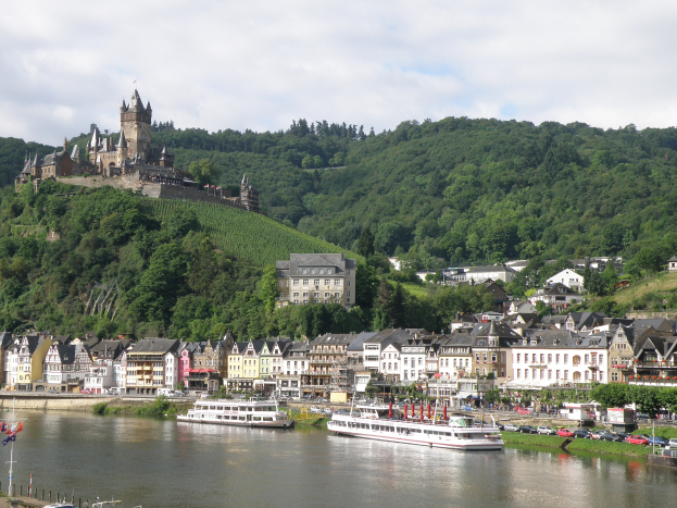 Ein malerischer Blick auf den Rhein in Deutschland mit einer Burg auf einem Hügel, Booten auf dem Fluss, Fahrzeugen auf einer nahen Straße und einer bewölkten Himmel.