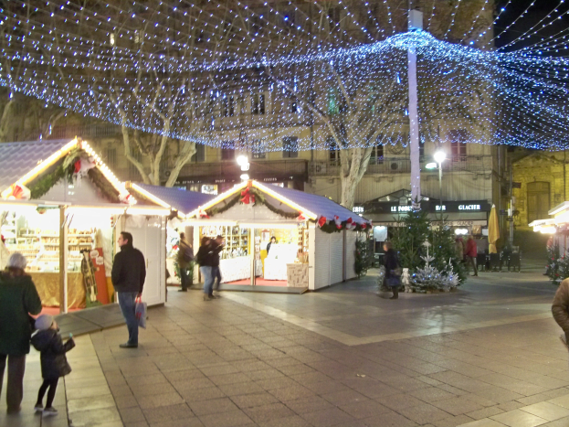 Ein lebendiger Weihnachtsmarkt in der Nacht in einer Stadtmitte, mit Menschen, die gehen, Ständen, die mit Lichtern geschmückt sind, und Gebäuden, Bäumen und Laternen im Hintergrund unter einem dunklen Himmel.