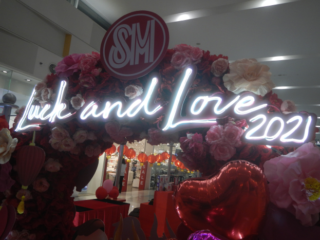 Ein Geschäft mit einem "Glück und Liebe 2021"-Schild, umgeben von Blumen, Ballons und Dekorationen, mit anderen Geschäften, Lichtern und einer Wand im Hintergrund, die eine festliche Atmosphäre für eine Valentinstag-Feier schaffen.