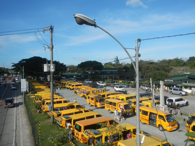 Eine große Anzahl gelber Schulbusse, die auf der Seite einer Straße geparkt sind, mit Menschen, die auf dem Gehweg gehen, Strommasten mit Drähten, Laternen, Bäumen, Gebäuden und einem bewölkten Himmel im Hintergrund.