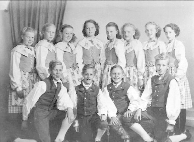 Ein Schwarz-Weiß-Foto einer Gruppe von Kindern, die vor einer Wand mit einem Vorhang posieren, mit dem Text "1930er-Kinder-Schule" unten.