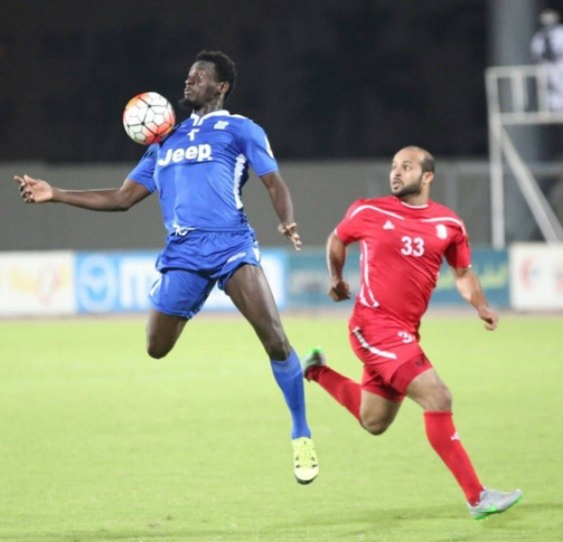 Zwei Männer beim Fußballspielen auf einem Feld, einer trägt ein blaues T-Shirt und Shorts, der andere ein rotes Kleid, im Hintergrund sind Bretter mit Text, ein Geländer, ein Pfosten und stehende Menschen zu sehen.