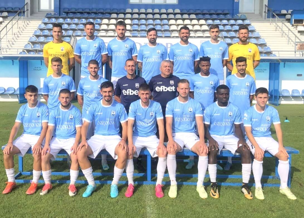 Eine Gruppe von Männern, einige sitzen auf Bänken und andere stehen auf einem Fußballfeld, mit Stühlen, Treppen, Geländern, Wänden und Fenstern im Hintergrund, wahrscheinlich feiern sie einen Mannschaftssieg.