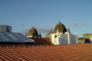 Stadtpanorama mit Gebäuden im Vordergrund unter einem blauen Himmel, mit Solarpanelen auf einem Dach, die den Einsatz erneuerbarer Energien anzeigen.