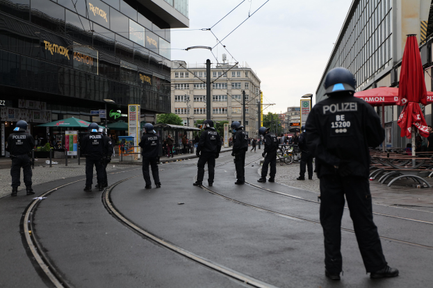 Polizeibeamte in schwarzer Uniform und Helm auf einer Straße mit Gebäuden, Bäumen und verstreuten Gegenständen im Hintergrund.