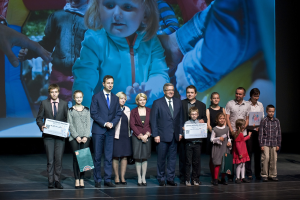 Eine Gruppe von Menschen auf einer Bühne mit Taschen und Urkunden, im Hintergrund ein Bildschirm mit einem Kind und anderen Menschen.