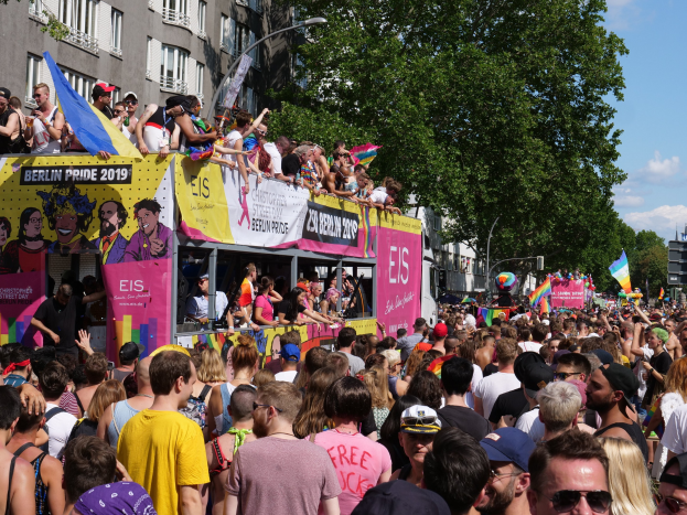 Eine große Menge marschiert auf der Straße während des Christopher Street Day 2019 in Berlin, viele tragen Mützen und Schutzbrillen, einige halten Fahnen, mit Bannern, Laternenpfählen, Bäumen und Gebäuden im Hintergrund unter einem bewölkten Himmel.
