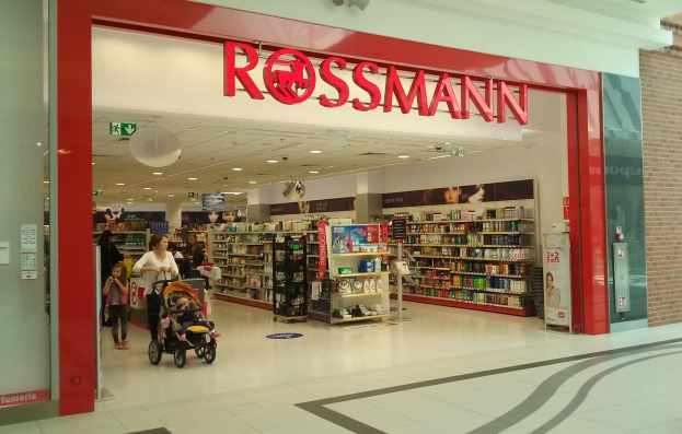 Ein Rossmann-Laden in einem Einkaufszentrum mit Menschen, einer Person mit einem Kinderwagen und Regalen mit Waren unter Deckenleuchten und Schildern.