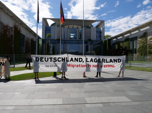 Gruppe von Menschen, die eine Fahne mit der Aufschrift "Deutschland, Lagerland Migration ist kein Verbrechen" vor einem Gebäude mit Fenstern halten, umgeben von Gras, einem Metallzaun, Pflanzen, Bäumen und Fahnenmasten bei einem bewölkten Himmel.