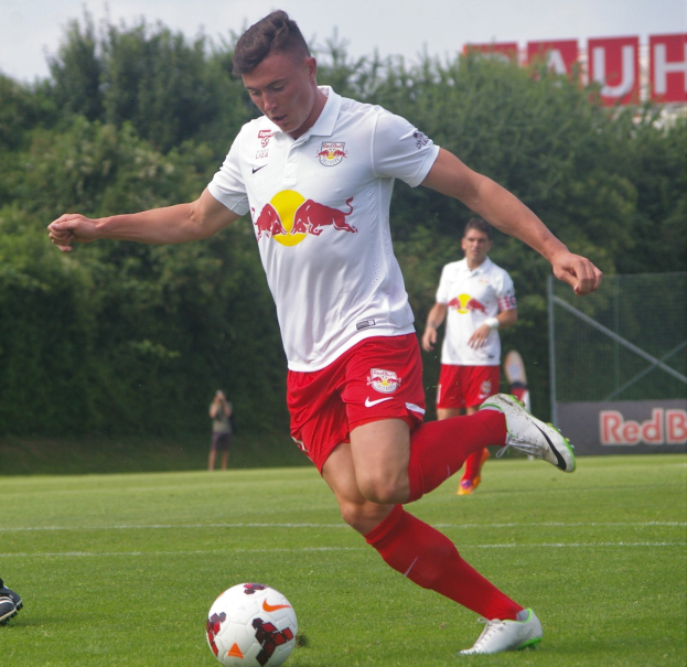 Ein Mann in einem Füßballtrikot schießt einen Ball auf einem grünen Feld mit Bäumen und einem klaren blauen Himmel im Hintergrund, mit einem Schild mit der Aufschrift "RB Leipzig" im Bild.