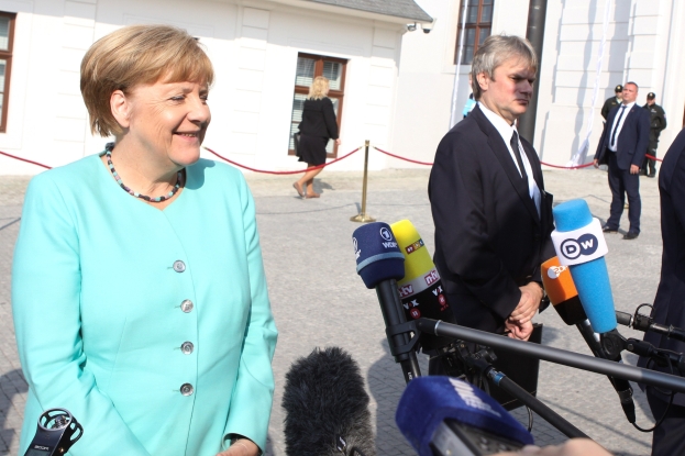 Bundeskanzlerin Angela Merkel beim Sprechen mit Reportern vor einem Gebäude mit bewölktem Himmel, umgeben von Menschen und Mikrofonen mit Ständern.