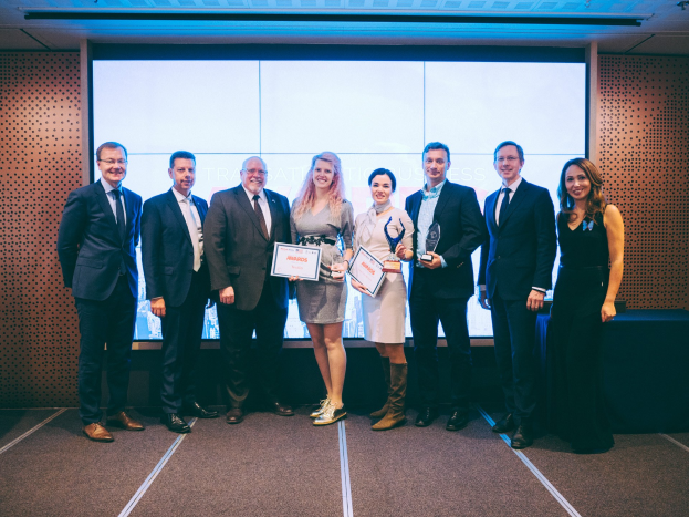 Eine Gruppe von Menschen, die Zertifikate halten und vor einem Bildschirm stehen, mit einem Tisch und einem Objekt an der Wand im Hintergrund, Gewinner der International Business Awards 2018.