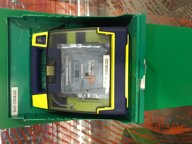 Eine grüne Box mit einem gelben und blauen Defibrillator-Gerät darin, das auf einem Tisch steht.