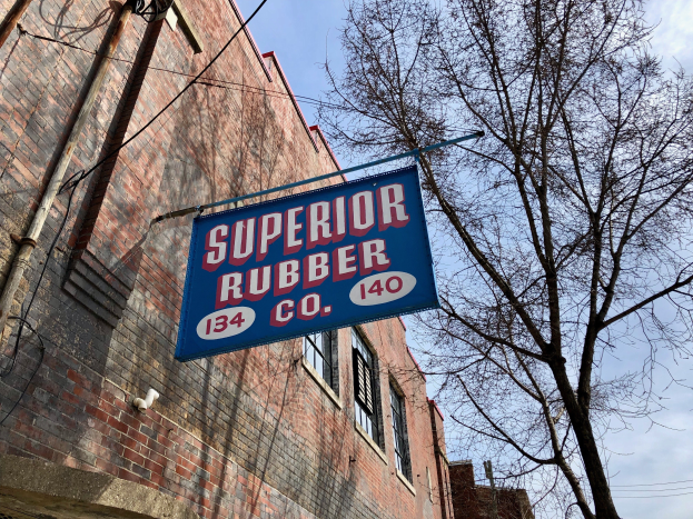 Ein Gebäude mit Fenstern, Rohren und einem Schild mit der Aufschrift "Superior Rubber Co" ist zu sehen, rechts daneben ein Baum und ein klarer blauer Himmel im Hintergrund.