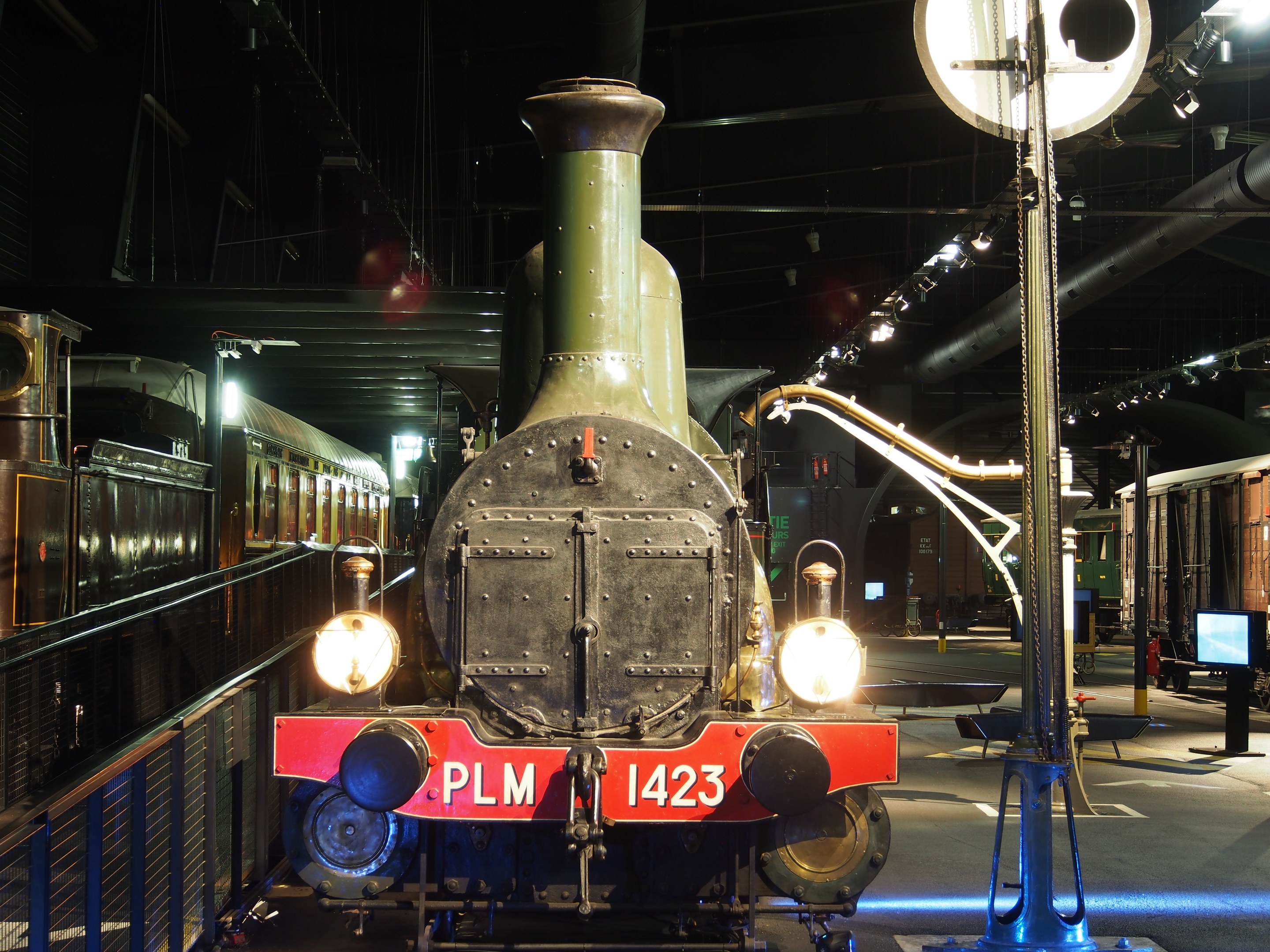 Dampflokomotive in einem Museum ausgestellt, umgeben von einem Metallzaun, mit Besuchern, Beschilderung, Beleuchtung und strukturellen Elementen.