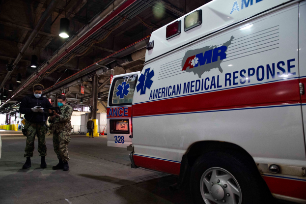 Ein Notarztwagen der American Medical Response (AMR) steht in einem Gebäude, vor ihm stehen zwei maskierte Personen, links ein Fahrzeug und im Hintergrund sind mehrere Personen, Lichter und Rohre zu sehen.