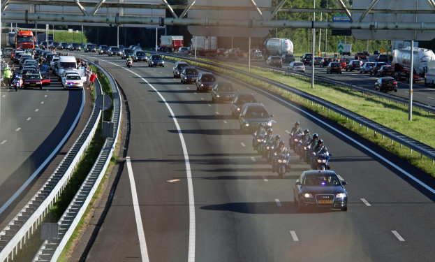 Autobahn mit dichtem Verkehr aus Autos und Motorrädern, gesäumt von Geländern und Gras, mit Bäumen, Polen, Texttafeln und einer Brücke mit Stangen im Hintergrund.
