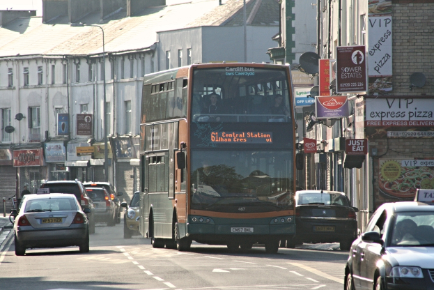 Eine Straße mit Autos und einem Bus im Vordergrund, mit Gebäuden mit Wänden, Fenstern, Tellern und Dächern im Hintergrund sowie Plakaten und Bannern an den Wänden und einem Laternenmast.