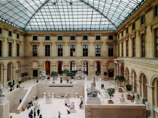 Großes gläsernes Dach des Louvre-Museums in Paris mit Statuen, Topfpflanzen, einer Treppe, Säulen, Fenstern und einem Metalldach mit Besuchern auf dem Boden.