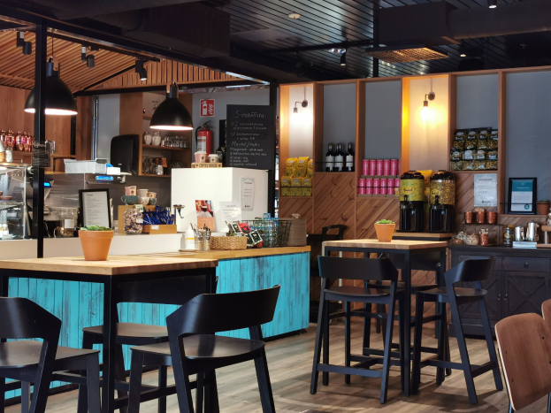 Innenraum eines Restaurants mit Holztischen, Stühlen, einem Blumentopf mit einer Pflanze auf einem Tisch, einer Schiefertafel, einem Kühlschrank, einem Feuerlöscher, Regalen mit Flaschen, Schränken mit Gegenständen, einem gerahmten Foto an der Wand und Deckenleuchten.