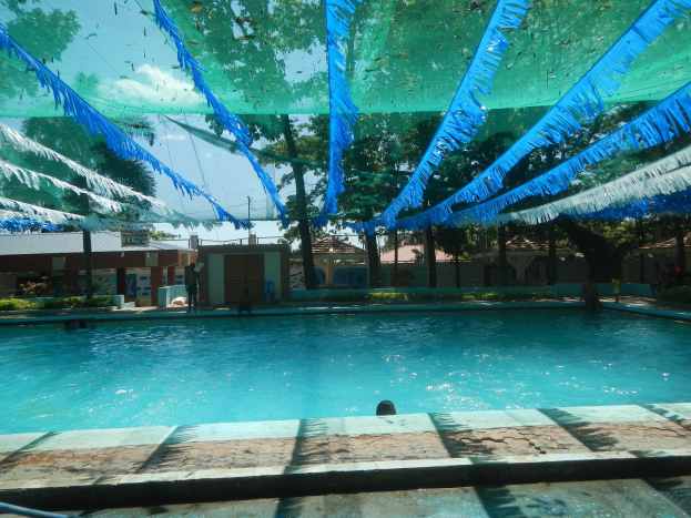 Großer Swimmingpool mit blauen und grünen Bändern an der Decke, umgeben von Häusern, Bäumen und einem klaren blauen Himmel, mit Menschen im Wasser.