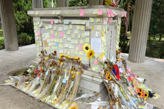 Ein Gedenkstein in einer grünen Fläche umgeben von Bäumen und Pflanzen, geschmückt mit Post-it-Zetteln und Blumen.