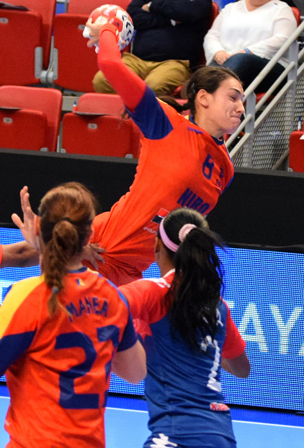 Eine Gruppe von Frauen, die Handball in einem Stadion spielt, mit einer, die den Ball hält, und Zuschauern im Hintergrund.
