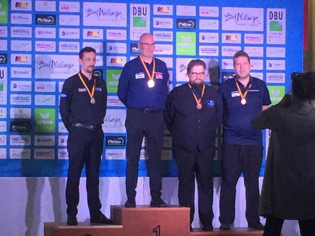 Fünf Männer auf dem Treppchen, ein Fotograf hält den Moment fest, im Hintergrund eine "DDBU Weltmeisterschaft 2019"-Plakette.