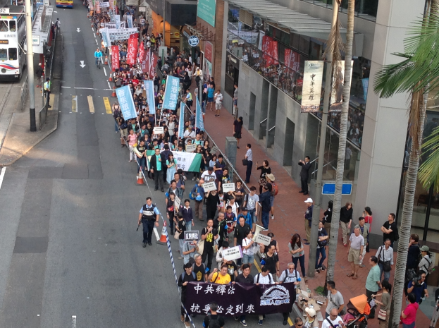 Eine große Gruppe von Menschen marschiert auf einer Straße in Hong Kong, mit Schildern und Transparenten, mit Bäumen, glaswandigen Gebäuden, Fahrzeugen und Schildern auf beiden Seiten.