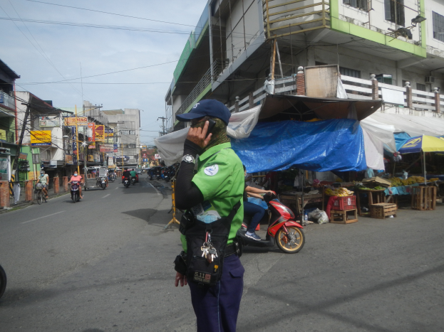 Ein Polizeibeamter in grüner und blauer Uniform und blauer Mütze steht an der Seite einer Straße, spricht in ein Handy, mit Fahrzeugen im Hintergrund, Ständen zu seiner Rechten und einem bewölkten Himmel darüber.