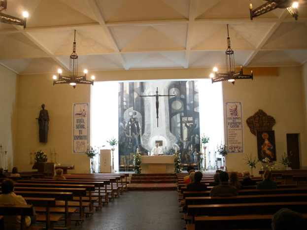 Eine Kircheninnenansicht mit Menschen auf Kirchenbänken, einer Person an einem Rednerpult mit Mikrofon, Kronleuchtern, Blumensträußen, Kerzen und Plakaten an den Wänden.