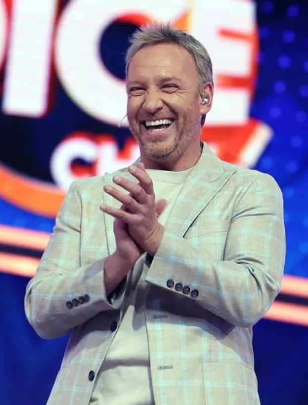 Ein Mann im Anzug steht auf einer Bühne, lächelt und klatscht, mit einem Schild 'The Voice' im Hintergrund.