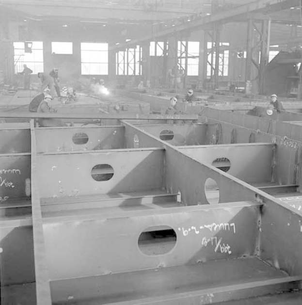 Ein Schwarz-Weiß-Foto einer Fabrik mit Arbeitern in Helmen, die an Stahlträgern arbeiten, mit Fenstern, Polen, Lichtern im Hintergrund und Text unten.