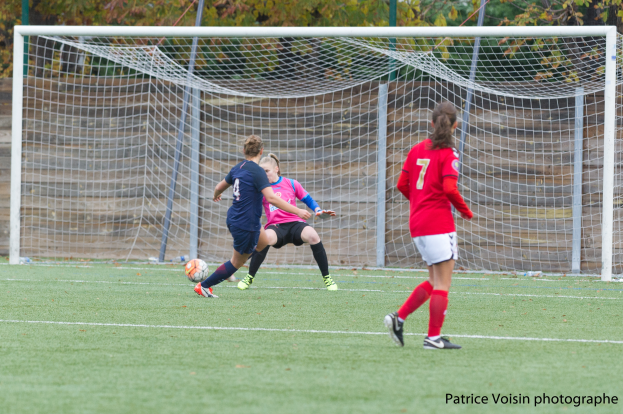 Eine Gruppe von Frauen, die auf einem grasbewachsenen Feld Fussball spielen, umgeben von Bäumen und mit einem Tor im Hintergrund sowie Text am unteren Bildrand.