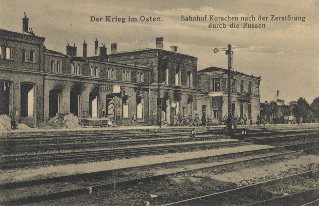 Altes Schwarz-Weiß-Foto des Bahnhofs Korschen in Ostern, Deutschland, zeigt einen Bahnhof mit Gleisen, Gebäuden, Pfählen, Menschen, Bäumen und einem Himmel im Hintergrund.