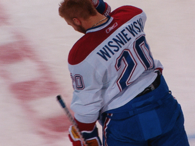 Ein Eishockeyspieler in einem "Wisniewski"-Trikot steht auf dem Eis und hält einen Eishockeyschläger, mit einem konzentrierten und entschlossenen Gesichtsausdruck.