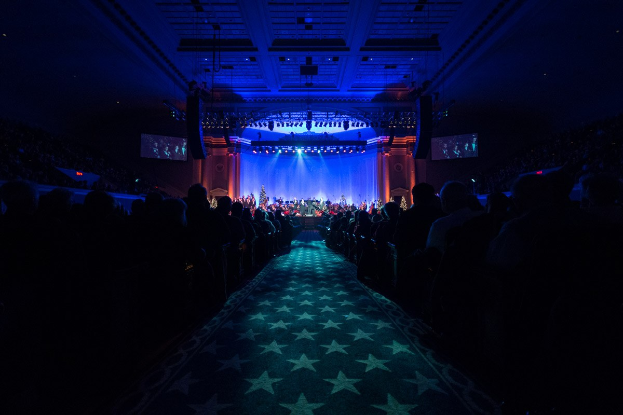 Ein großer Saal mit Sitzplätzen, eine Bühne mit Musikern und Deckenbeleuchtung und -fernseher, was auf ein Konzerthaus während der Nationalkonvention 2019 hindeutet.