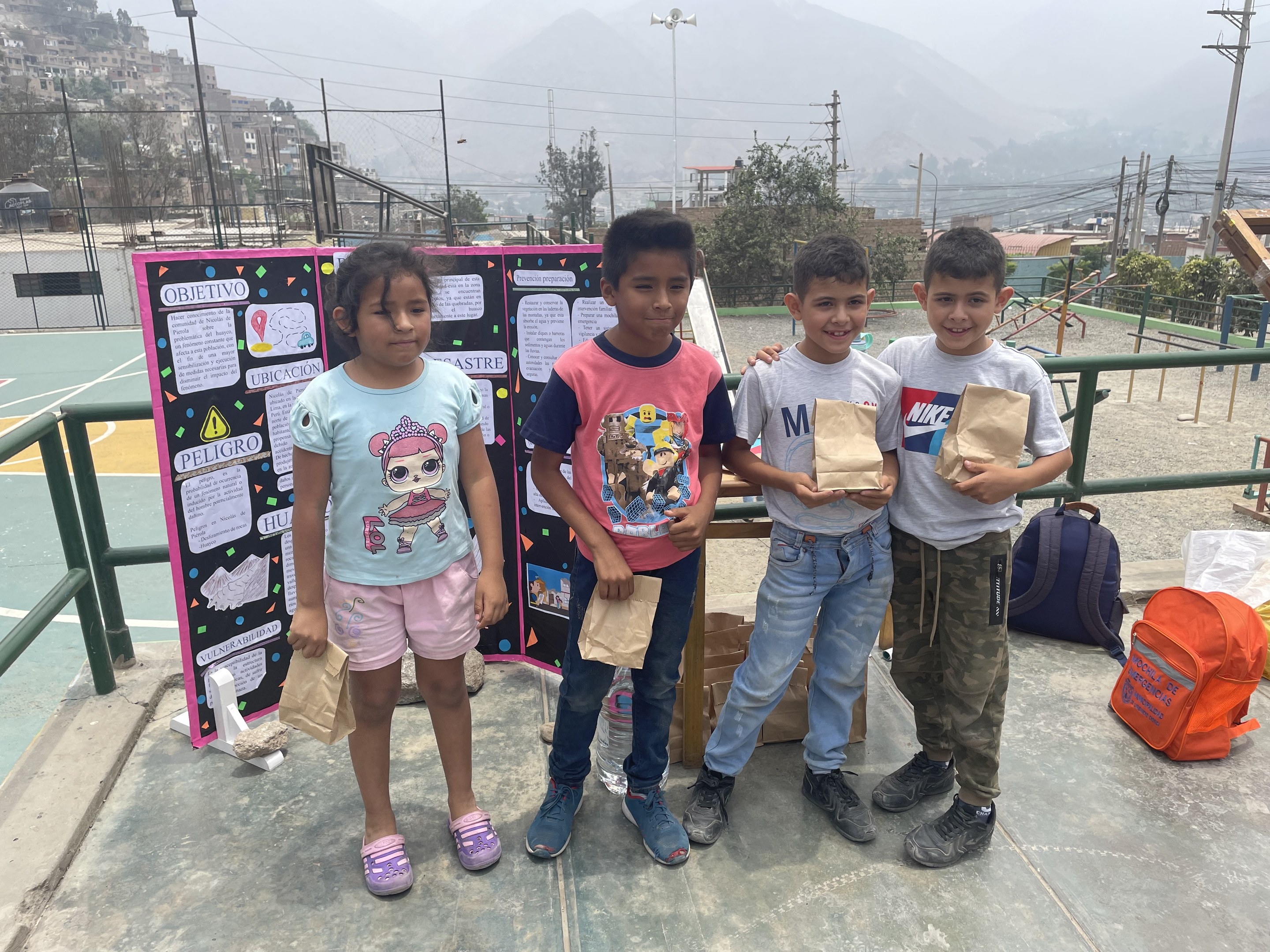 Gruppe von Kindern auf einem Basketballfeld mit Papiertüten in den Händen, Tüten und Gegenstände rechts und eine Tafel links, vor einem Hintergrund aus Zaun, Pfosten, Straßenlaternen, Bäumen, Gebäuden, Hügeln und Himmel.