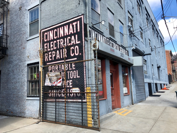 Das Cincinnati Electrical Repair Co. Gebäude mit Fenstern, Türen, einem Schild, einem Metallzaun, einem Müllbehälter, Pfählen, Drähten, Bäumen und einem bewölkten Himmel.