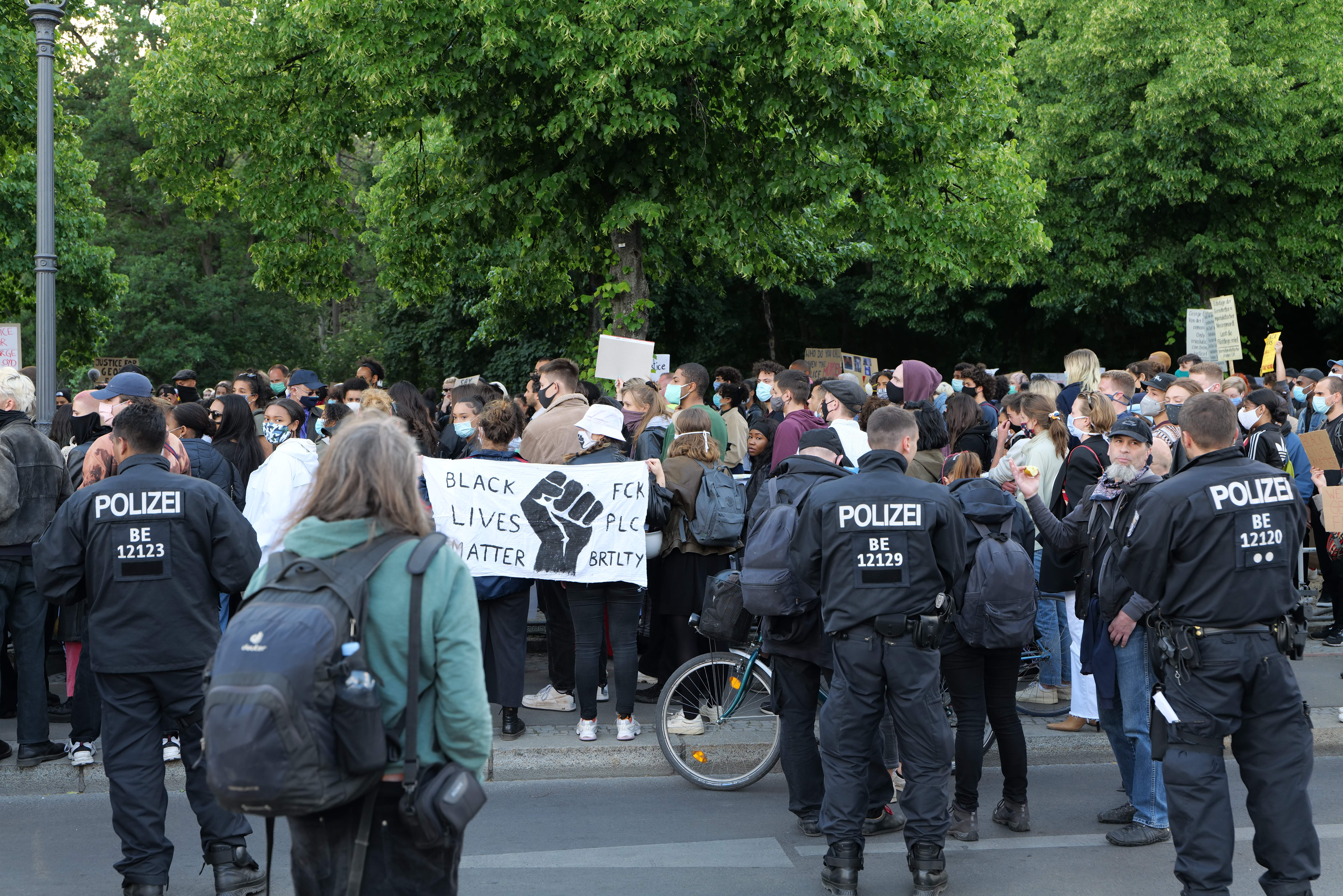 Eine große Gruppe von Menschen steht an der Straße, einige halten Schilder, andere tragen Kappen und Taschen, mit einem Fahrrad im Vordergrund und Bäumen und einem Pfahl im Hintergrund, bei einer Black-Lives-Matter-Demonstration in Berlin.