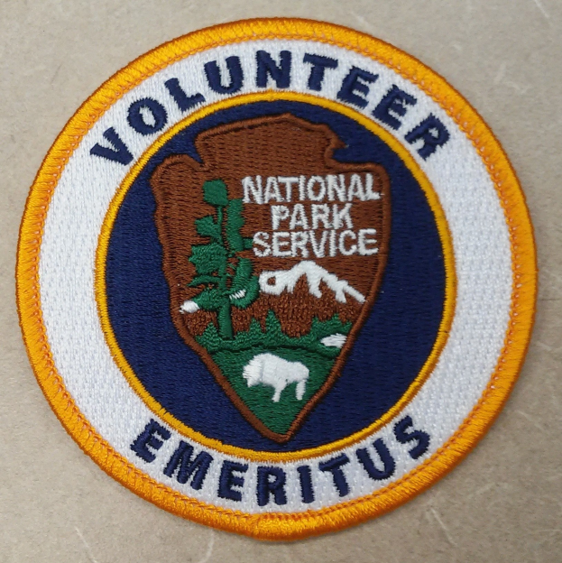 Runder Aufnäher mit "Volunteer National Park Service Emeritus" in fetter schwarzer Schrift auf einem weißen Hintergrund, umrahmt von einem blauen Rand und einem gelben Kreis.