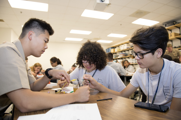 Three students at a table in a classroom work on a project with papers, pens, a mobile phone, and other items; racks with objects, ceiling lights, and a projector are in the background.