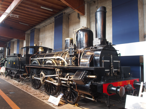 Eine große, alte Dampflokomotive auf Eisenbahnschienen in einem Museum ausgestellt, umgeben von Informationsschildern, Ständen und anderen Ausstellungsstücken, mit einer beleuchteten Decke und Wand im Hintergrund.