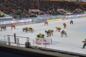 Gruppe von Menschen, die Hockey auf einem Eisplatz mit Netz und Umzäunung spielen, Zuschauern im Hintergrund, Banner sichtbar, Stadionbeleuchtung oben.